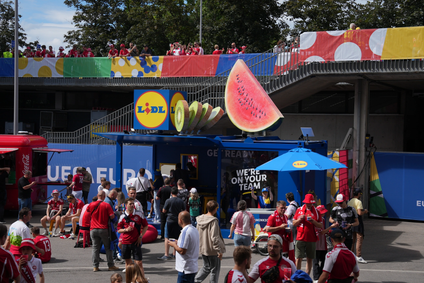 Fußballfans versammeln sich vor einem blauen Lidl-Verkaufsstand mit riesigen Obst-Attrappen während der UEFA EURO.