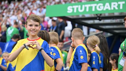 A smiling boy in a yellow, blue, and red jersey stands in the foreground of a football stadium, making a heart shape with his hands. In the background, other children in similar jerseys and a crowded spectator stand are visible beneath a sign that reads "STUTTGART".