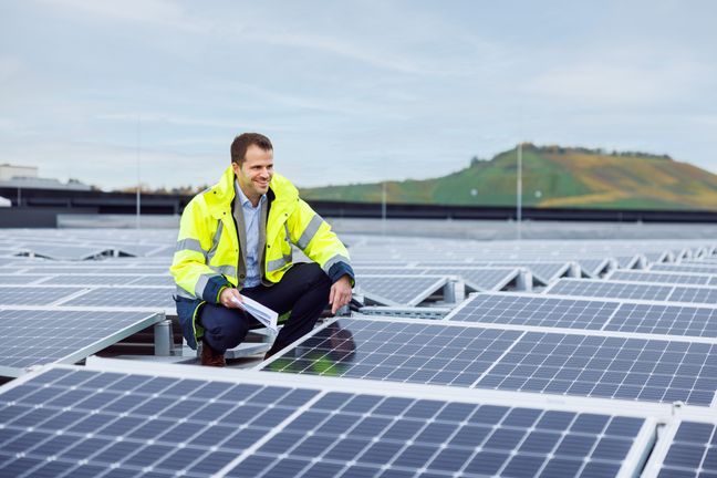Net Zero durch Photovoltaik: Ein lächelnder Experte in Warnschutzjacke begutachtet Solarmodule auf einem Industriedach.