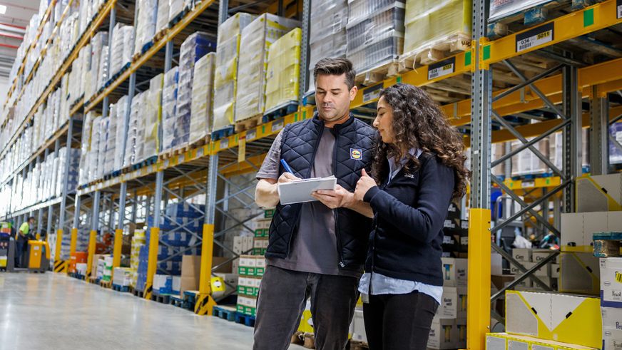 Zwei Lidl-Logistik-Mitarbeiter prüfen konzentriert Bestände in einem modernen, hell beleuchteten Palettenlager.