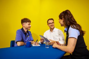 Two Lidl employees and an applicant in a shirt engaged in conversation in a relaxed atmosphere at a blue table.