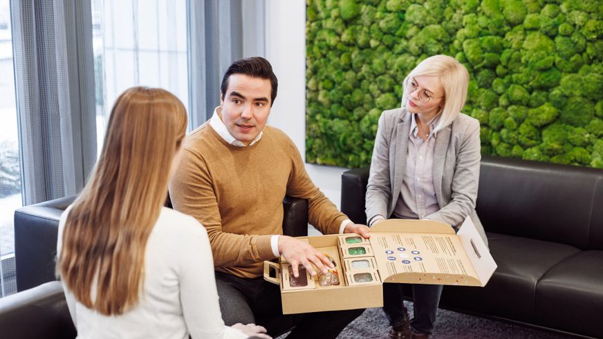 Ein Berater präsentiert zwei Kundinnen in einem modernen Büro eine Musterbox mit nachhaltigen Verpackungsmaterialien.