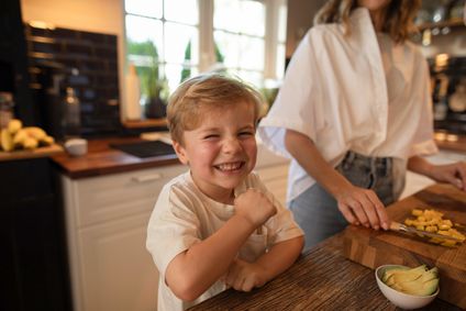 Ein strahlender kleiner Junge freut sich am Tisch in einer hellen Küche über die Zubereitung von frischem Obst und Avocado.