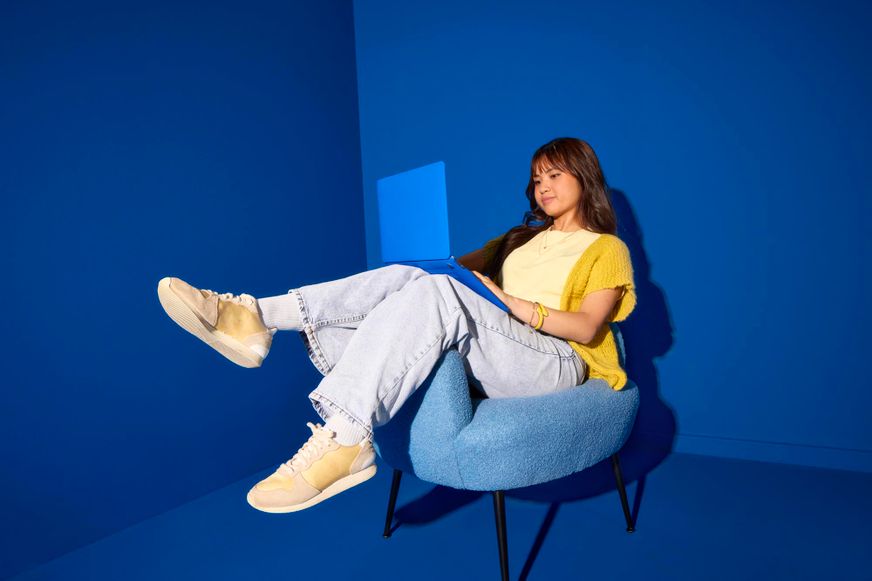 Lässig in einem blauen Sessel liegend, arbeitet eine junge Frau im gelben Cardigan mit ihrem Laptop in einem gänzlichen blauen Raum.