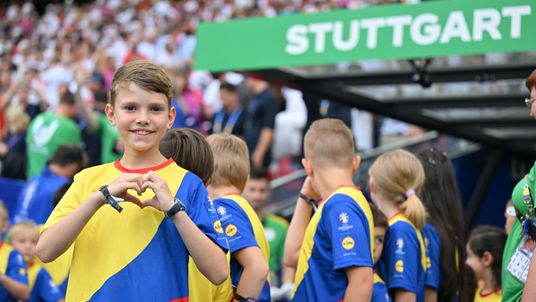 A smiling boy in a yellow, blue, and red jersey stands in the foreground of a football stadium, making a heart shape with his hands. In the background, other children in similar jerseys and a crowded spectator stand are visible beneath a sign that reads "STUTTGART".
