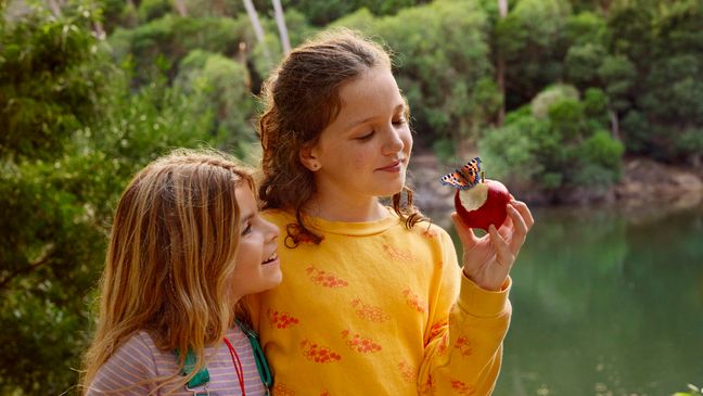 Zwei Mädchen stehen lächelnd am Seeufer, während ein Schmetterling auf dem angebissenen Apfel in der Hand eines Mädchens landet.