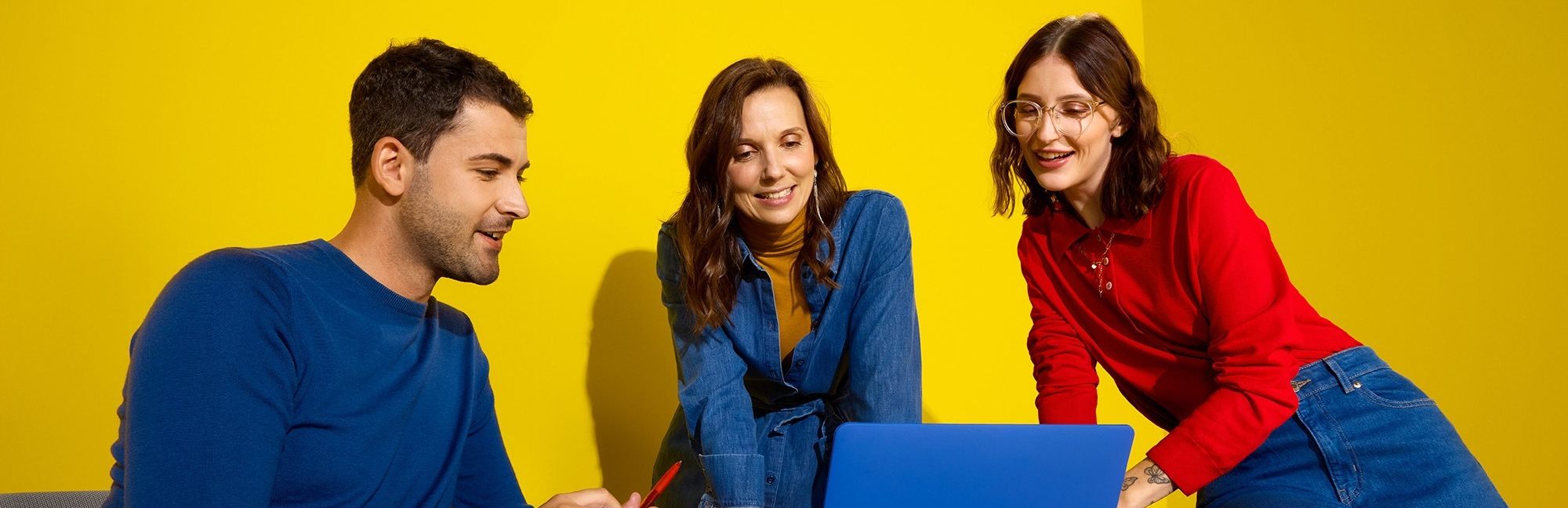 Drei lächelnde Lidl-Mitarbeitende blicken in einem modernen, gelben Büro Hintergrund gemeinsam auf einen blauen Laptop.
