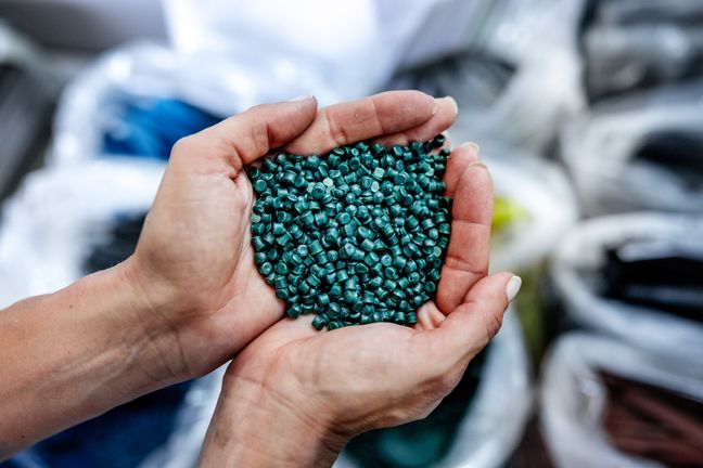 Zwei Hände halten eine große Menge dunkelgrünes Kunststoff-Recyclinggranulat in einer detaillierten Nahaufnahme.