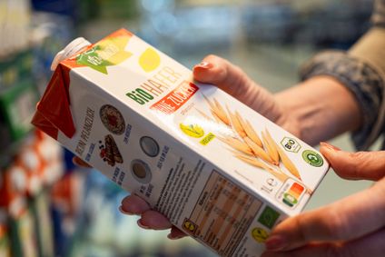 Nahaufnahme von Händen die ein Tetrapack Lidl Bio-Haferdrink ohne Zuckerzusatz vor einem Supermarktregal halten.