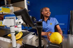 A warmly smiling Lidl cashier scanning a honeydew melon, embodying the fast and friendly service at Lidl checkouts.