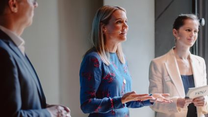 Fokus auf eine engagiert sprechende Sprecherin in Blau, die mit zwei Personen im Rahmen der Mitarbeiter Initiative YOU auf der Bühne steht.