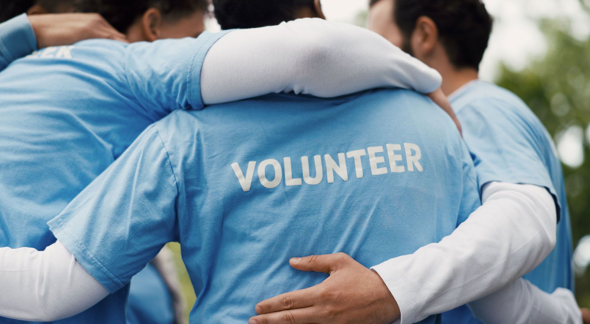 Gruppe von Freiwilligen in blauen T-Shirts steht eng umarmt zusammen; auf dem Rücken steht „Volunteer“.