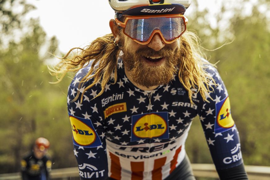 Nahaufnahme eines lächelnden Radrennfahrers mit langen Haaren, Bart und Sonnenbrille von Lidl-Trek beim Fahren im Regen.