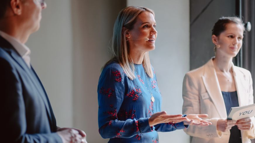 Fokus auf eine engagiert sprechende Sprecherin in Blau, die mit zwei Personen im Rahmen der Mitarbeiter Initiative YOU auf der Bühne steht.