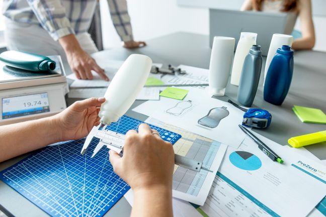 Hände vermessen eine Kunststoffflasche mit dem Messschieber zur präzisen Verpackungsoptimierung am Arbeitsplatz.