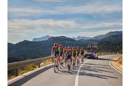 Eine Gruppe von Lidl-Trek Radsportlerinnen und Radsportlern fährt in Teambekleidung eine Bergstraße hinauf, gefolgt von einem Begleitfahrzeug vor einer Bergkulisse.