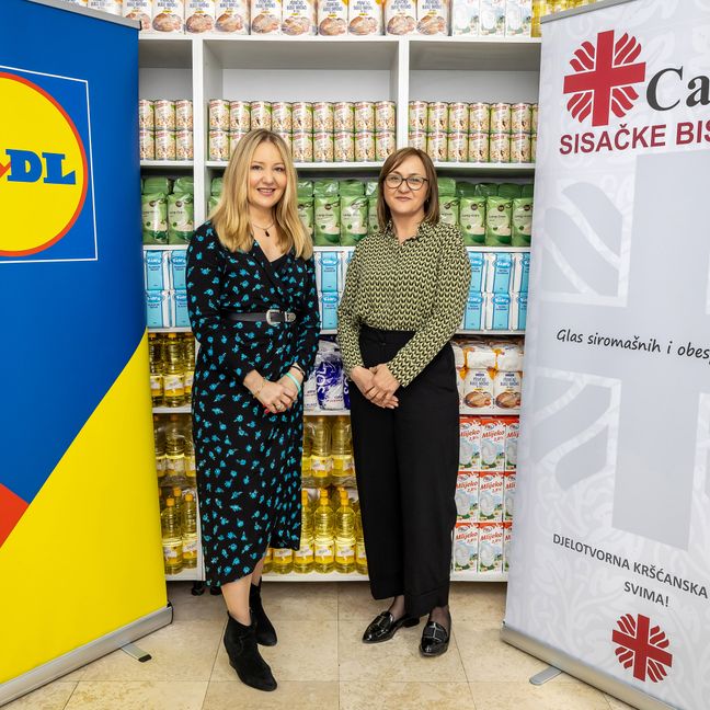 Marina Dijaković iz Lidla Hrvatska i Kristina Radić stoje između Lidl i Caritas banera ispred polica s prehrambenim donacijama.