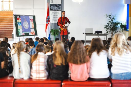Predavač u crvenom speleološkom odijelu drži kacigu i drži predavanje učenicima koji sjede u učionici