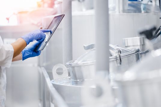 Eine Person in Schutzkleidung steht in einem hellen Labor und bedient ein Tablet zur digitalen Kontrolle.