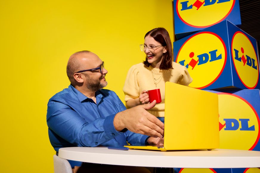 Dvoje nasmijanih kolega radi za stolom sa žutim prijenosnim računalom ispred Lidl pozadine.Dvoje nasmijanih kolega radi za stolom sa žutim prijenosnim računalom ispred Lidl pozadine.