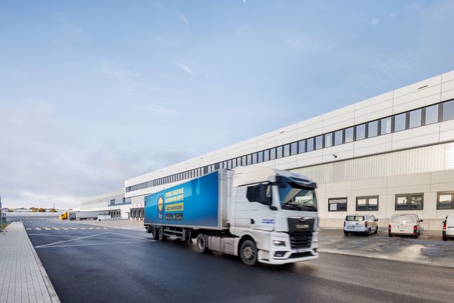 Ein Lidl E-Lkw fährt bei Tageslicht an einem modernen Logistikzentrum vorbei.