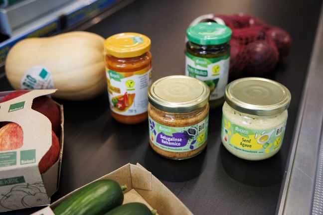 Verschiedene vegane Vemondo Bio-Aufstriche liegen neben frischem Kürbis und Äpfeln auf einem schwarzen Supermarkt-Kassenband.