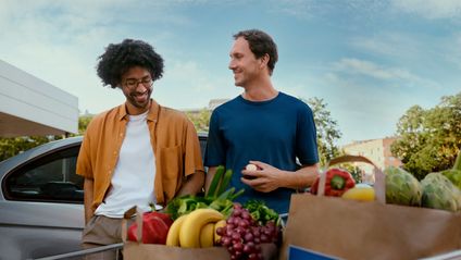 Zwei Männer unterhalten sich gut gelaunt an einem Auto vor prall gefüllten Einkaufstüten mit frischem Obst und Gemüse.