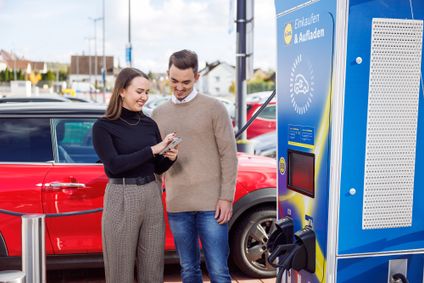 Ein lächelndes Paar bedient per Smartphone eine E-Ladestation auf einem Lidl Kundenparkplatz.